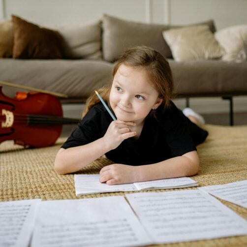 Girl lying on the floor writing in a book with a 'cello in the background